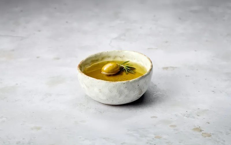 A small stoneware bowl of green olive oil with an olive and a sprig of rosemary.